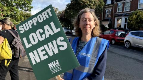 Woman with long brown hair holds green "support local news " placard