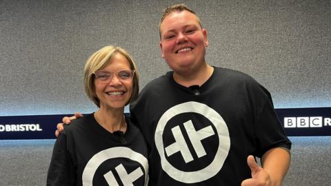 Denise Coles and Joe Sheppard. They are both wearing black t-shirts printed with a white Take That logo - two rotated Ts inside a circle. Joe has his arm around Denise and both of them are smiling at the camera