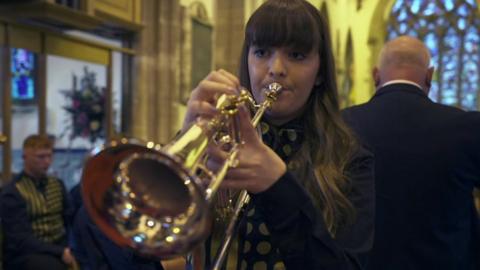 Teenage girl with long dark hair plays flugelhorn solo with band and conductor behind her