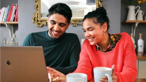 A young man and young woman sit in a living room with mugs of coffee looking at a laptop, smiling.