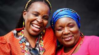 Tina Sekete (left) and Jane Dlamini smiling. They are wearing bright colours and have white make up on their face.