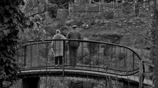 black and white image of two people on a bridge overlooking woodland