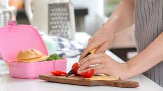 Mother cutting tomato for school lunch at table in kitchen (stock photo)