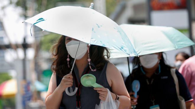 Duas mulheres usando ventiladores portáteis e guarda-chuvas enquanto caminham na rua