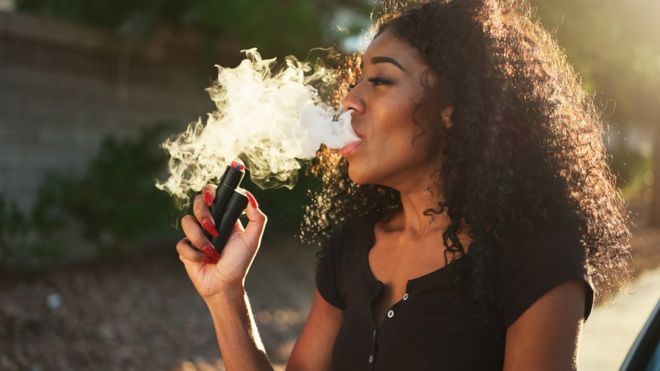 Une femme, vêtue d'un T-shirt noir, exhale un grand nuage de vapeur d'e-cigarette alors qu'elle fume à l'aide d'un vaporisateur.