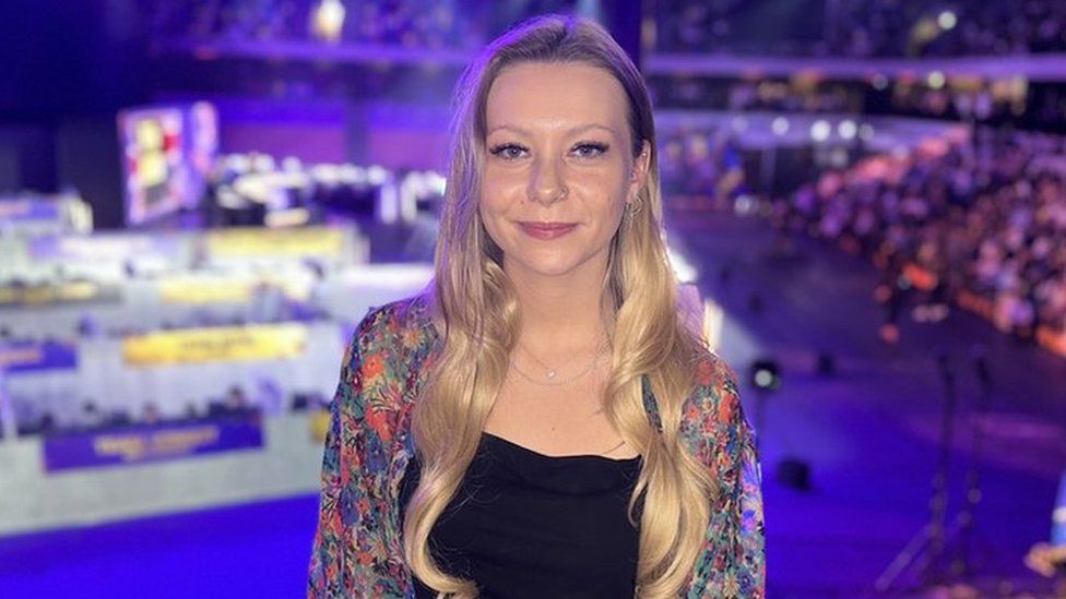 Kels stood smiling for the camera with the arena bathed in the purple glow of stage lights behind her in the background. She has long blonde hair worn down and a ringed piercing in her nose and right ear. She is wearing a black top with a multicoloured flowery patterned shirt over the top of it.