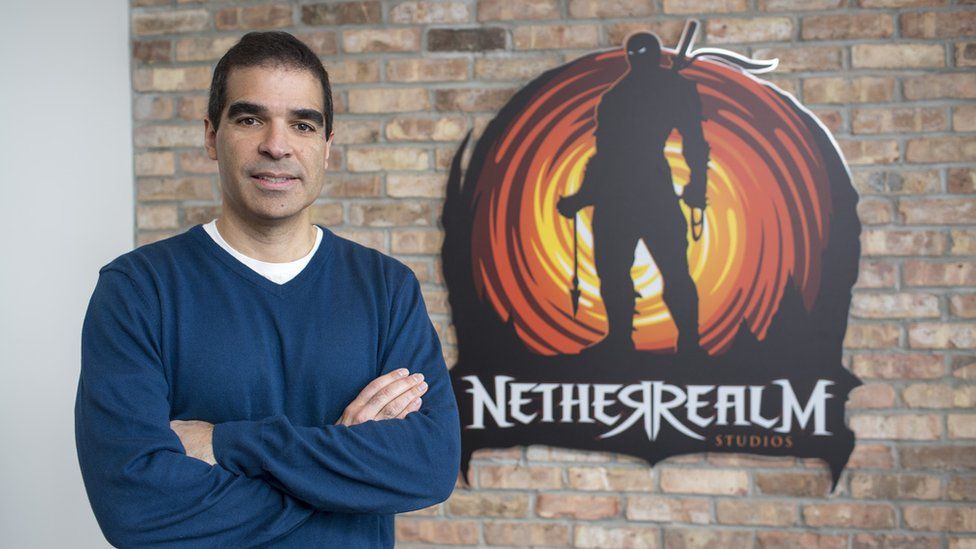 Ed Boon - a middle-aged man with short black hair - wears a navy v-necked sweater with white t-shirt underneath. He's standing, arms folded, in front of a brick wall with a sign that reads "NetherRealm Studios" on it. The sign shows a ninja character in silhouette, on top of a vortex design in red and yellow shades resembling a swirling fire