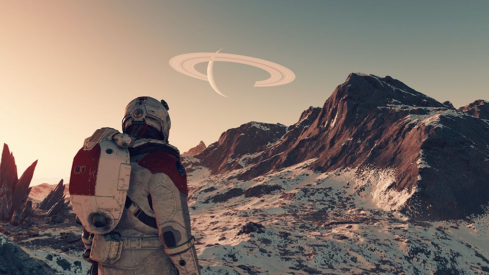 A screenshot from Starfield shows the back of an astronaut character in spacesuit and helmet surveying a barren planetscape. It is rocky, with cliffs jutting out of the ground. The rocks are covered with a white powder - either dust or snow. In the distance another planet, and its horizontal rings, can be see in the sky.