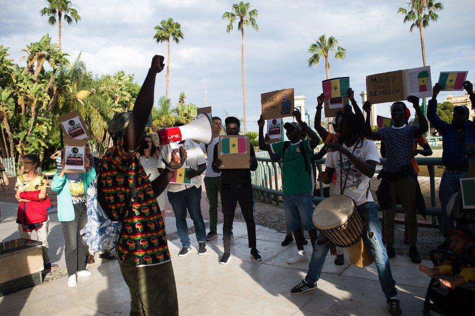 Senegalese protesters holding placards outside next to tall palm trees. One man is talking through a speakerphone and the other men appear animated - 7 June.