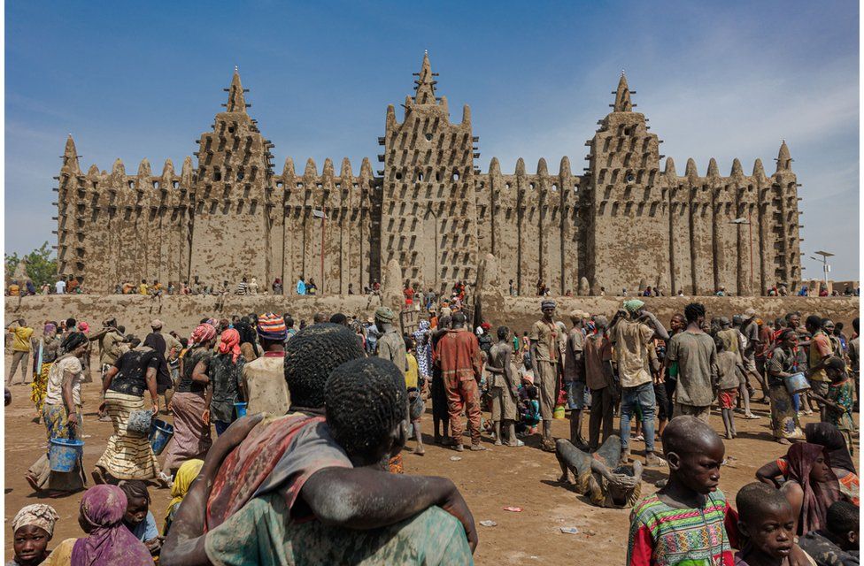 People staring at the Great Mosque of Djenne - 4 June. It is a large brown building which stands against a backdrop of blue skies.