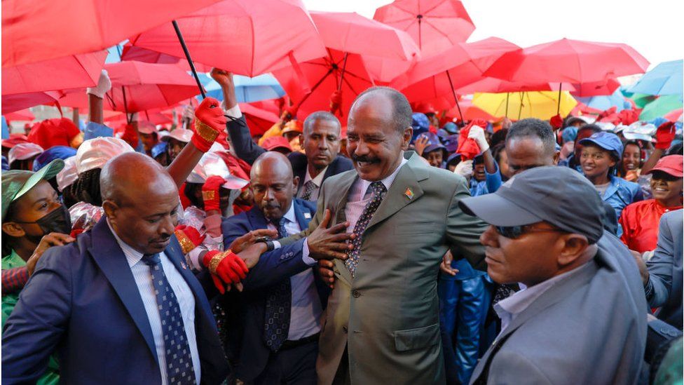 Eritrean president Isaias Afewerki surrounded by people holding red umbrellas. He is smiling.
