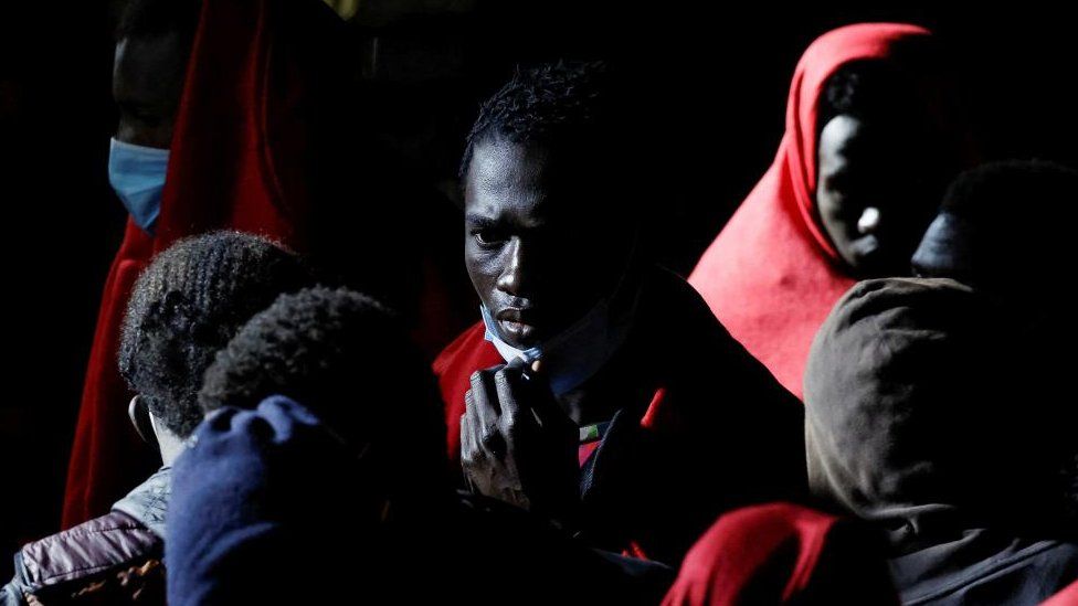 Migrants wait in a boat, one of whom holds a face mask below his chin